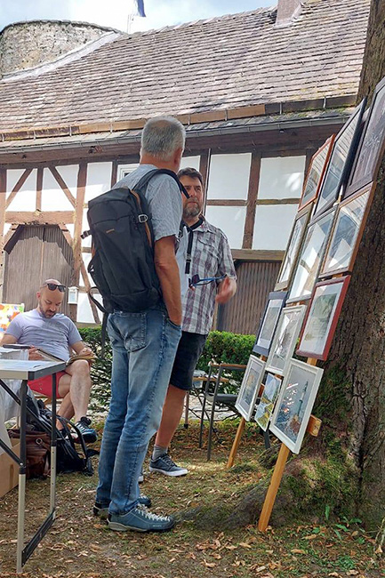 Besucher betrachten ausgestellte Kunstwerke und sprechen mit dem Künstler während der Aktion „Malen um die Burg 2025“ im Innenhof der Burg Dringenberg.